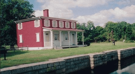 Waterfront view of a historic red building with white windows and white porch on green grass.