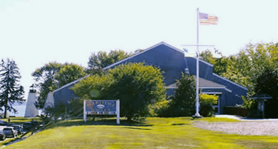 Blue triangular building with large green lawn and flagpole with lighthouse in the background.