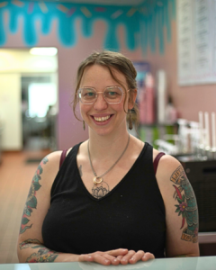 Bakery owner in Havre de Grace, Maryland, smiling in front of colorful wall behind treat counter
